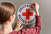 Eine Frau klebt einen großen runden DRK-Logo Aufkleber auf die Wand.