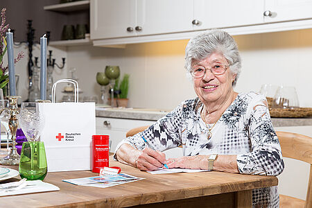 Ältere Frau sitz am Tisch und füllt ein Formular aus. Neben ihr steht die Rotkreuzdose.